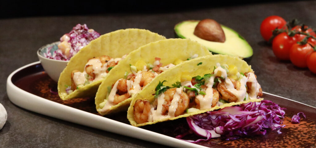 Shrimp tacos served in crispy corn tortillas topped with creamy sauce, fresh herbs, and mango salsa, plated with red cabbage, salsa, avocado, and cherry tomatoes.
